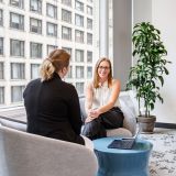 Two colleagues in conversation beside a tall office window, a laptop on the table between them