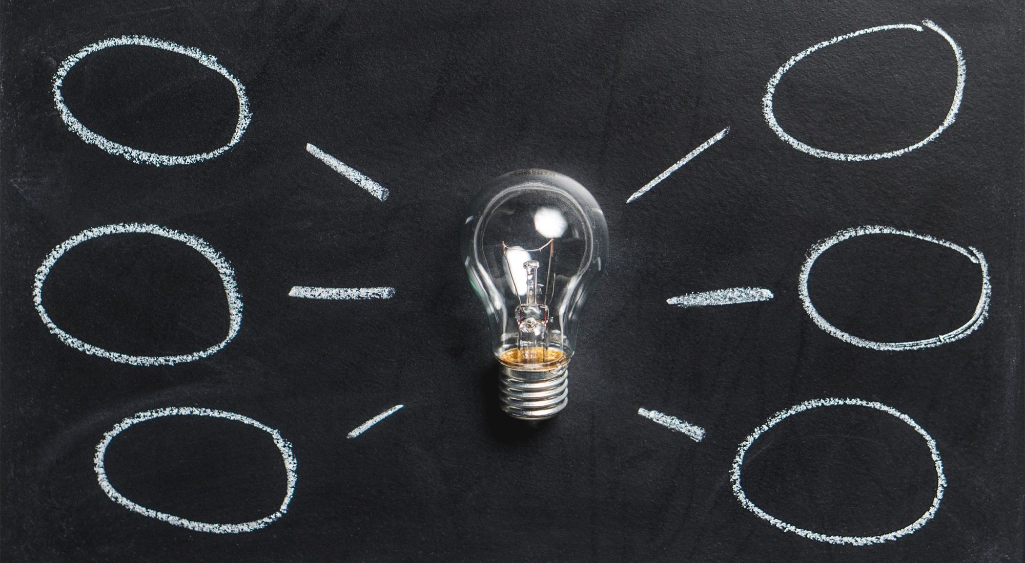 A lightbulb laying on a chalkboard with six idea bubbles drawn around it.