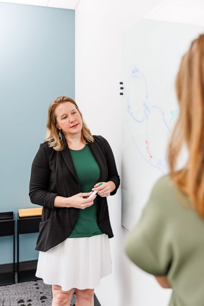 A facilitator holding a marker beside a whiteboard diagram, talking with a colleague
